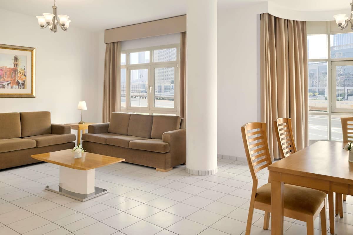 A modern living room with a sofa, a coffee table, a dining table with chairs, a chandelier, and large windows with curtains.