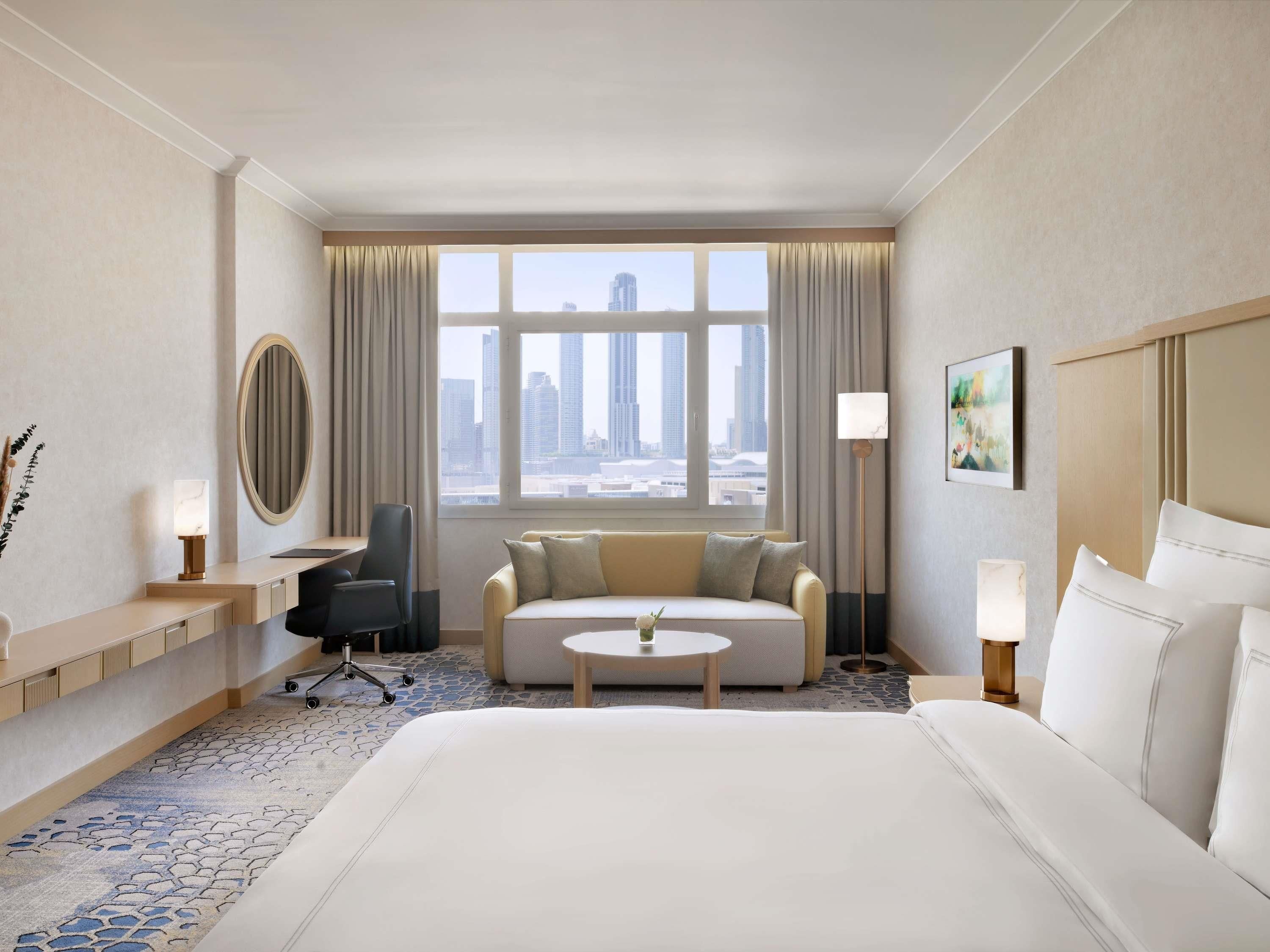 A hotel room with a bed, a sofa, a desk, a chair, a round table, a floor lamp, a painting, and a view of the cityscape.