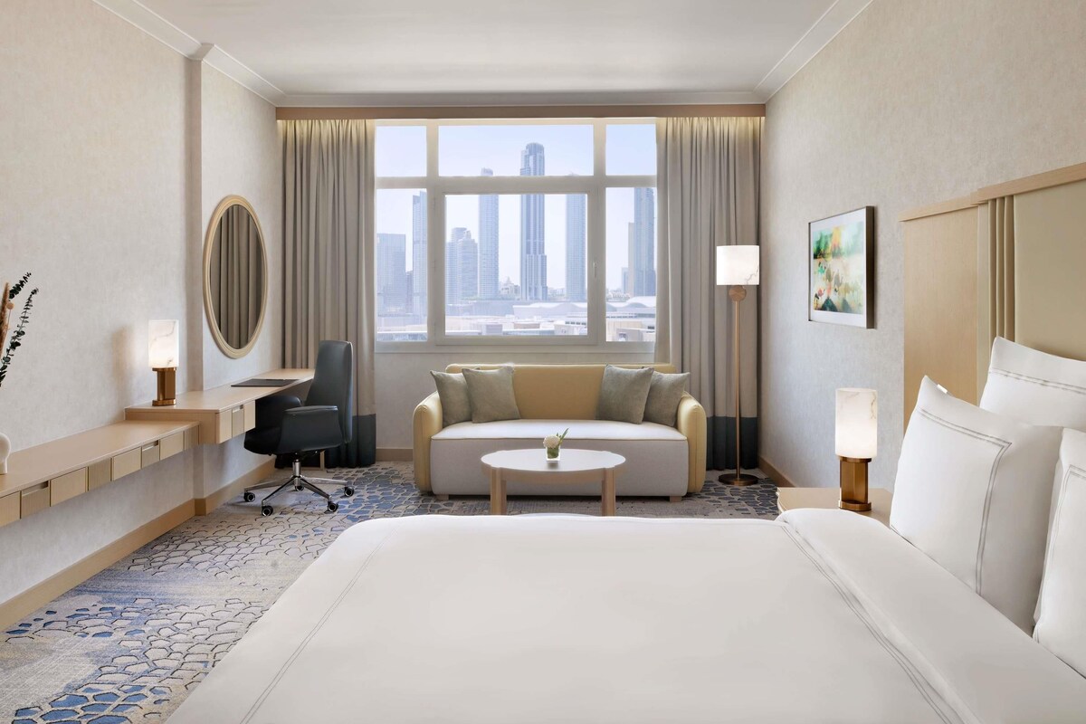 Habitación de hotel con cama, sofá, escritorio, silla, una mesa redonda, lámpara de pie, un cuadro y vistas a la ciudad.