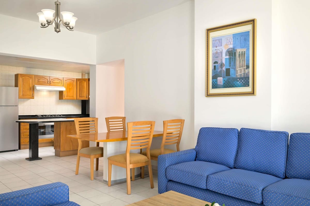 A living room with a blue sofa, wooden dining table, and chairs, a kitchen with wooden cabinets, and a framed picture on the wall.