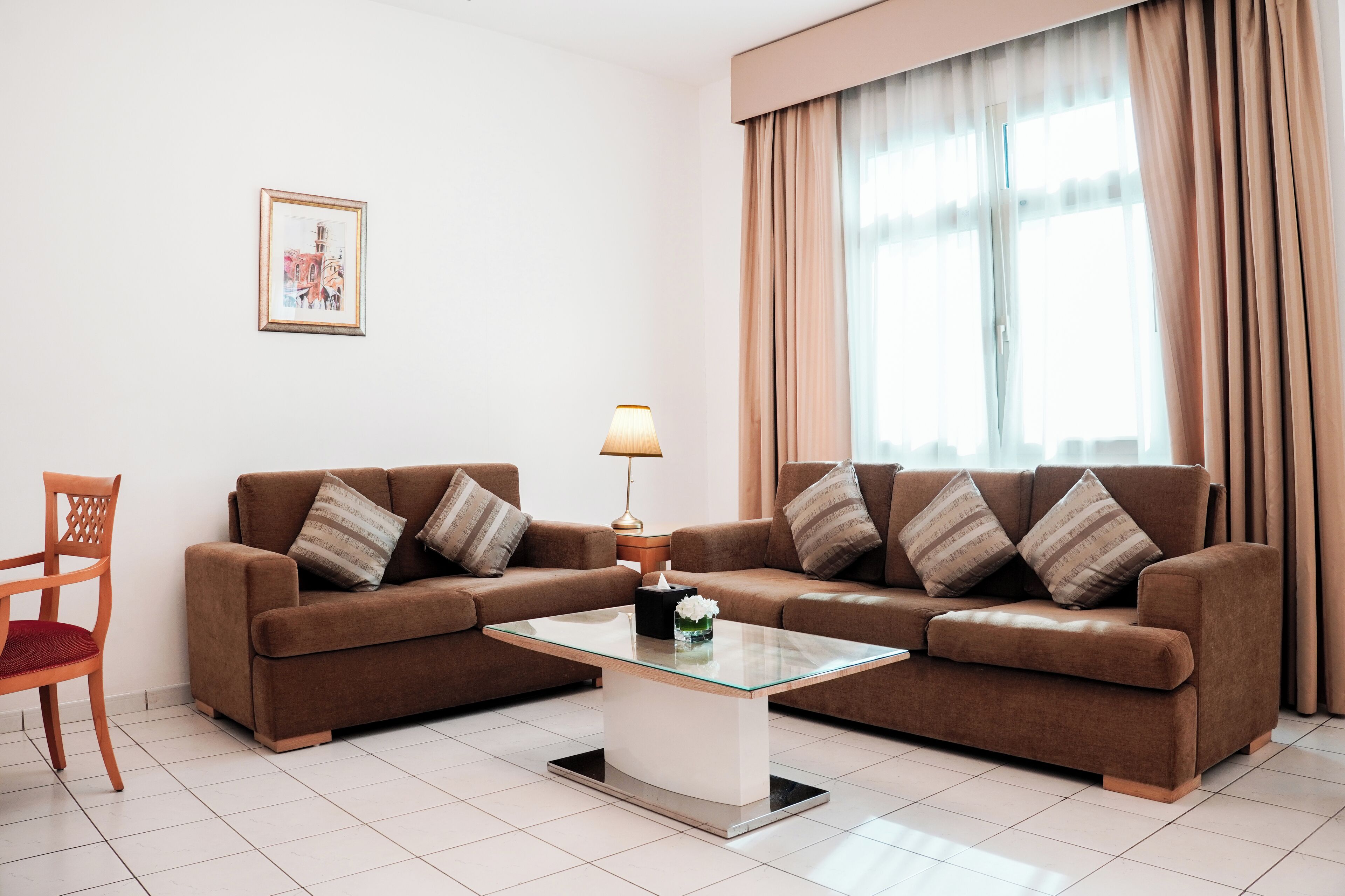 A living room with two brown sofas, a glass coffee table, a wooden chair, a framed picture on the wall, and a window with curtains.