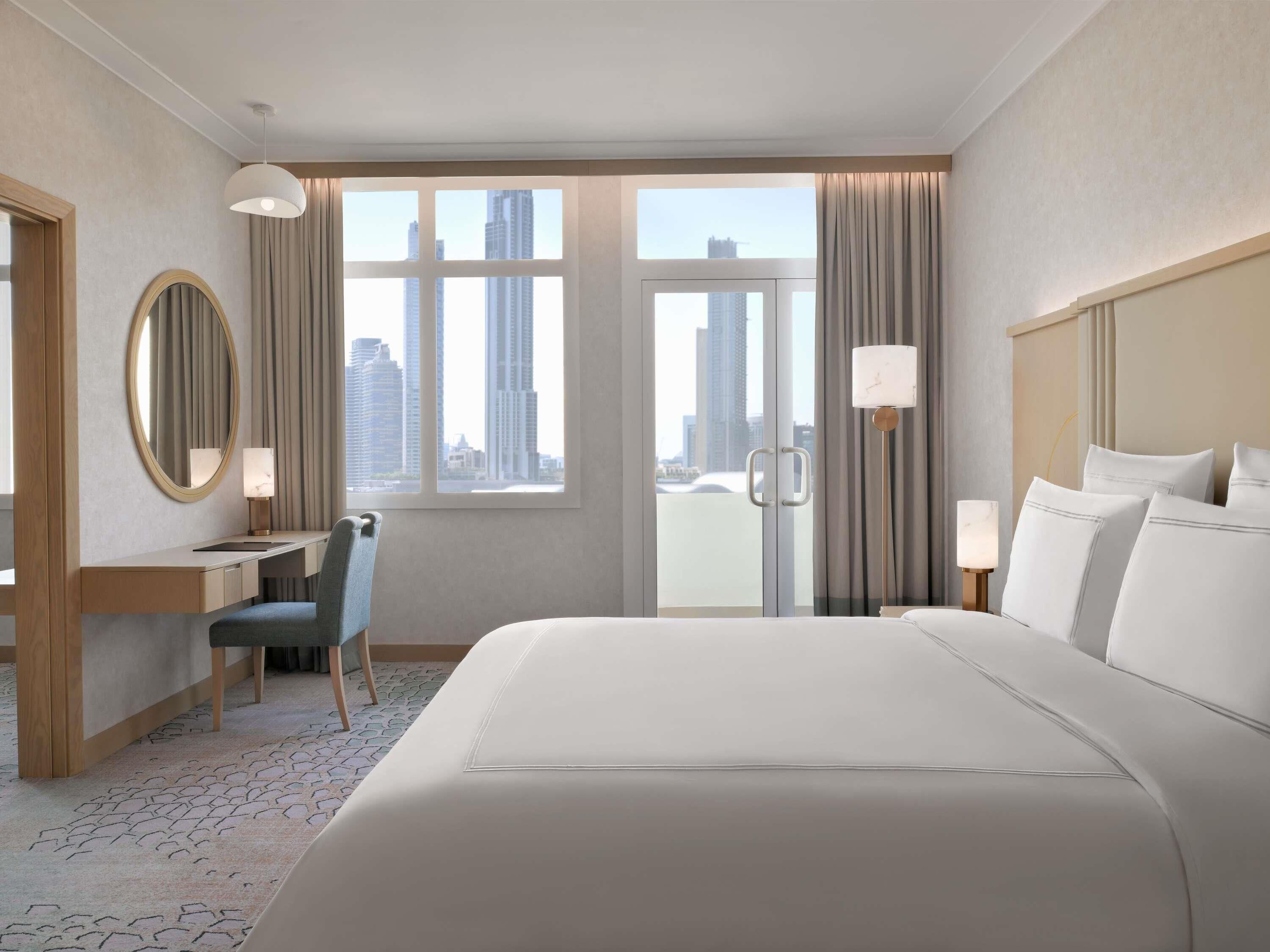 Habitación de hotel con una cama grande, un escritorio con silla, un espejo redondo y vistas a la ciudad a través de las ventanas.