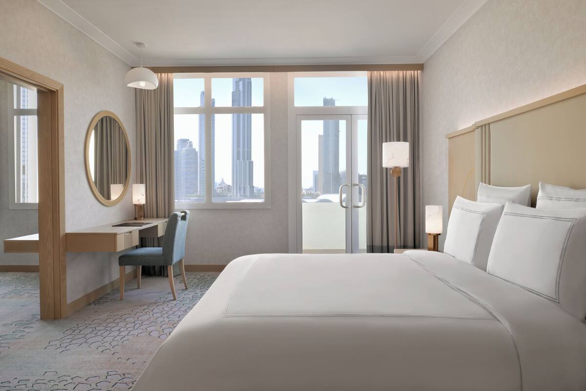 A hotel room with a large bed, a desk with a chair, a round mirror, and a view of tall buildings through the window.