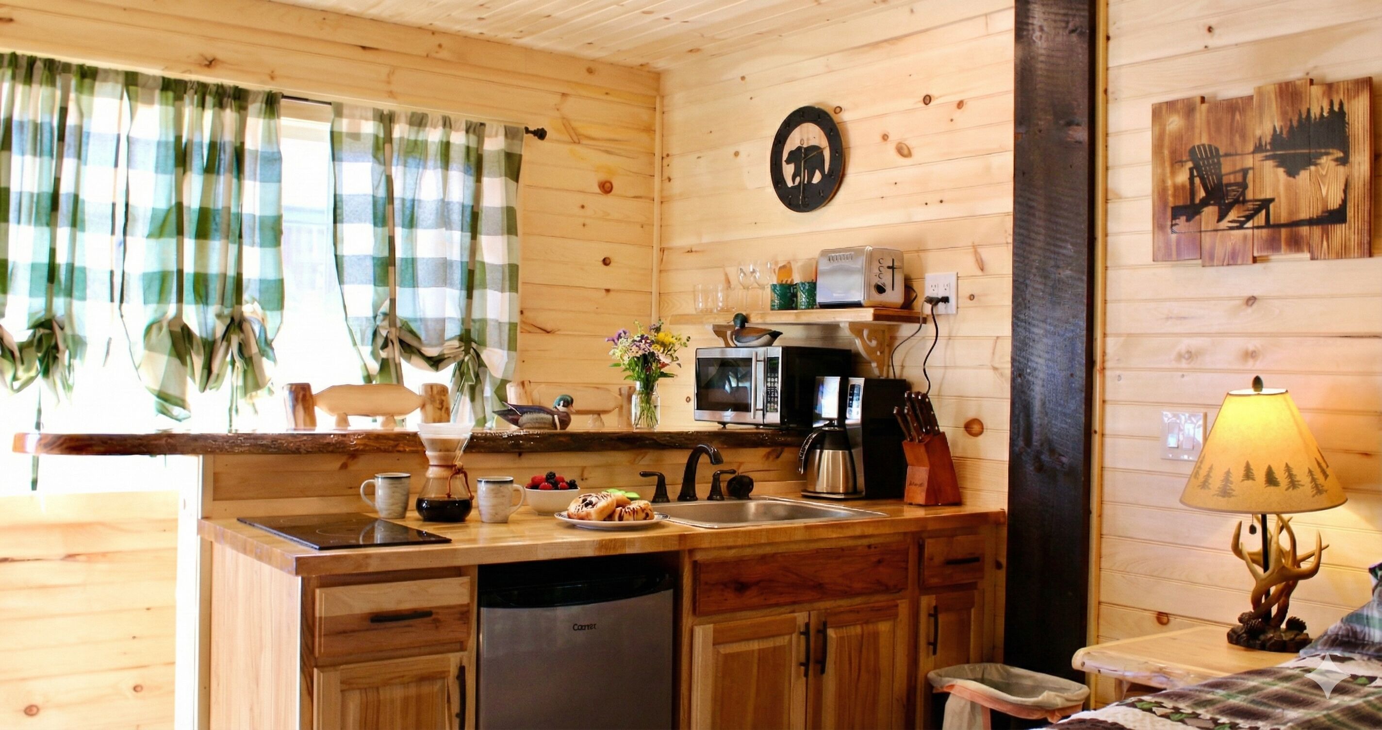 A wooden cabin interior with a kitchen area featuring a sink, stove, and microwave. There is a coffee maker, a toaster, and a lamp on the counter.