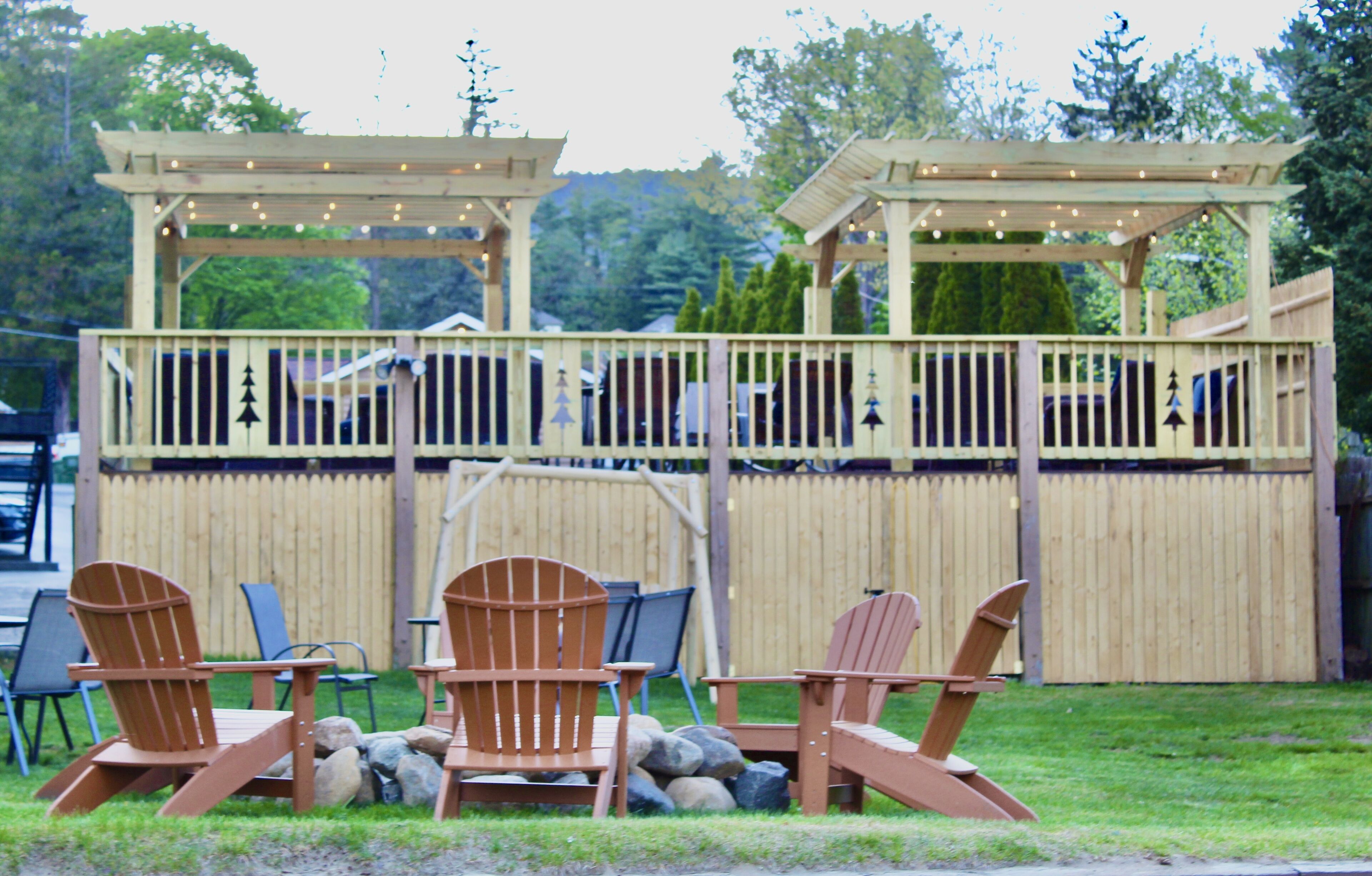 An outdoor seating area with wooden chairs and tables, a wooden pergola with string lights, and a wooden fence.