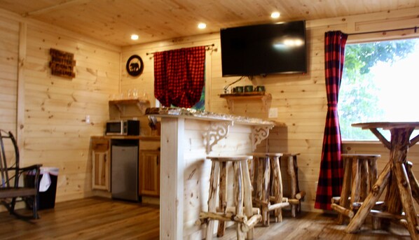 A cabin interior with wooden walls, a kitchen area, a TV mounted on the wall, and a window with curtains. - Sundowner on Lake George (Lake George)