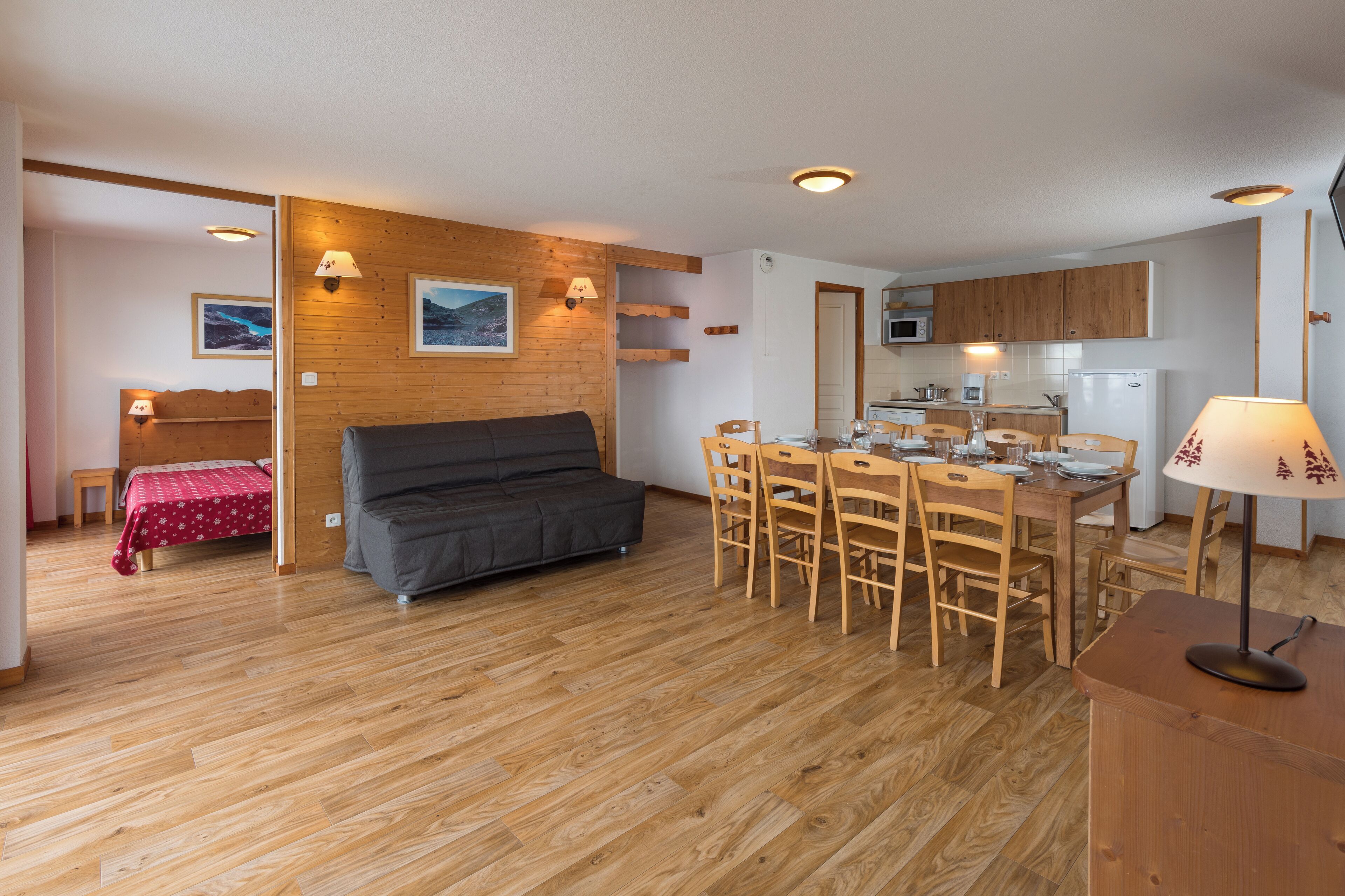 A compact living space with a wooden floor, a sofa, a dining table with chairs, a kitchen area, and a bed with a red bedspread.