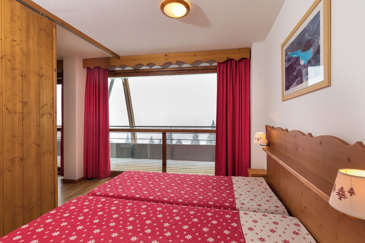 Une chambre avec un lit, une tête de lit en bois, une fenêtre avec des rideaux rouges et un tableau encadré au mur.