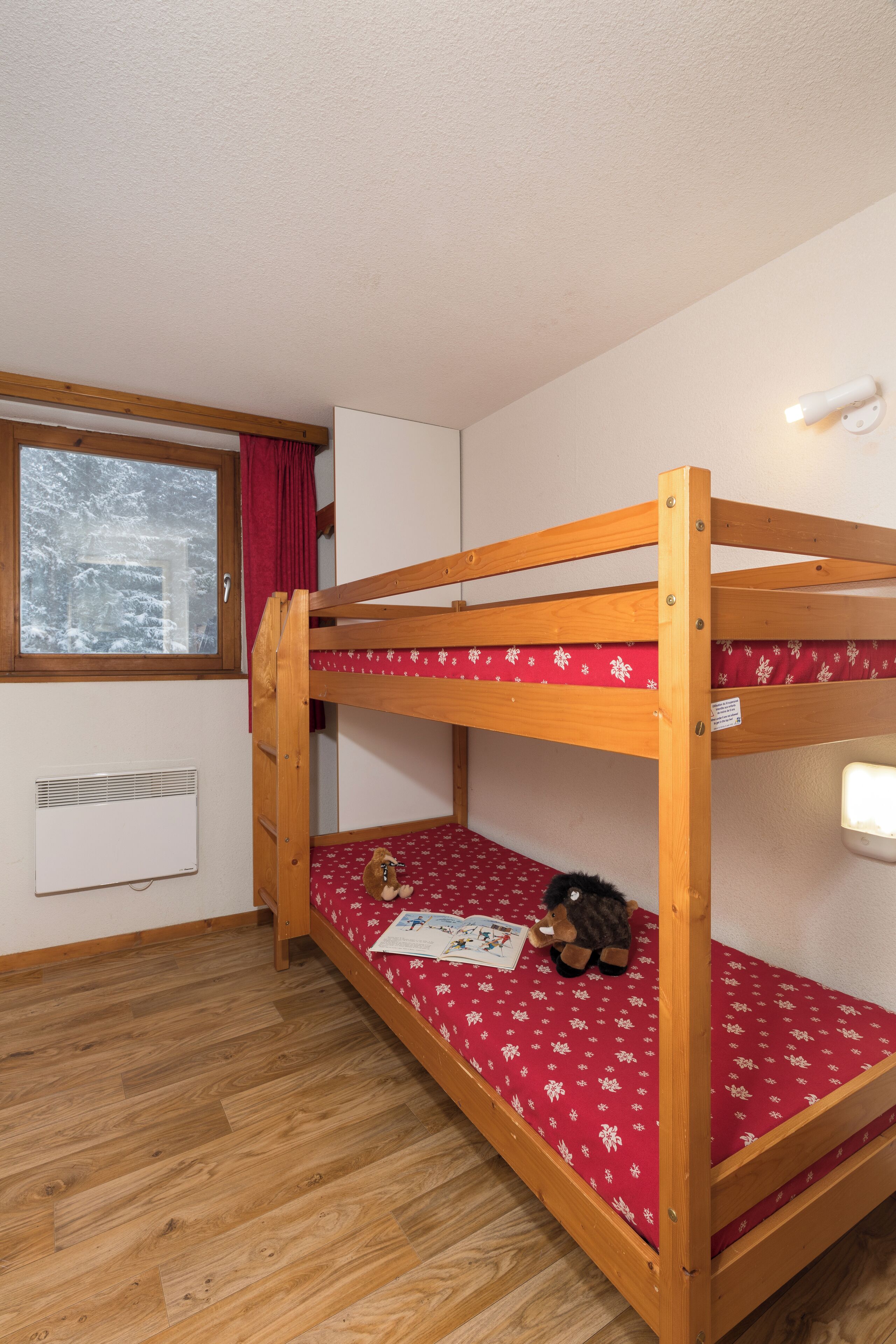 A bunk bed with a red mattress, a window with a view of snow-covered trees, and a white wall with a thermostat.