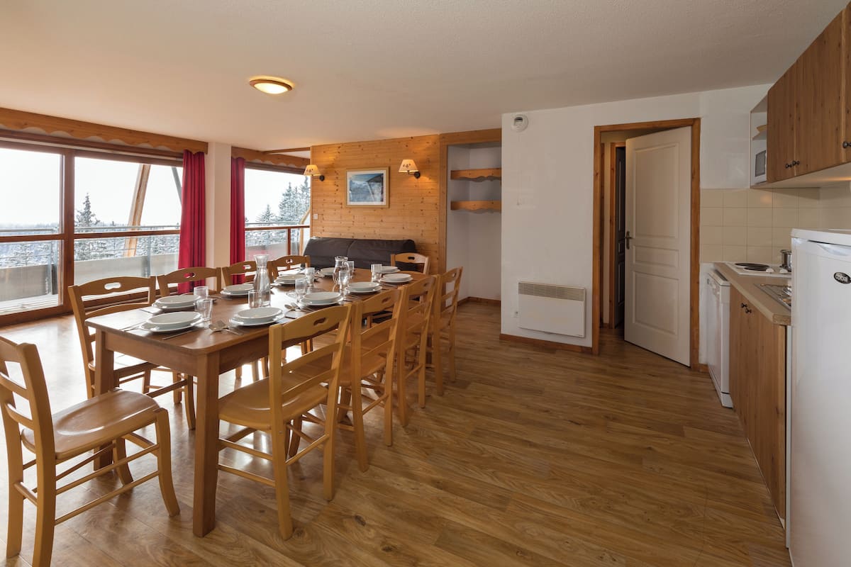 Un coin repas avec une table en bois dressée pour quatre personnes, des chaises en bois et une vue sur un paysage enneigé par de grandes fenêtres.