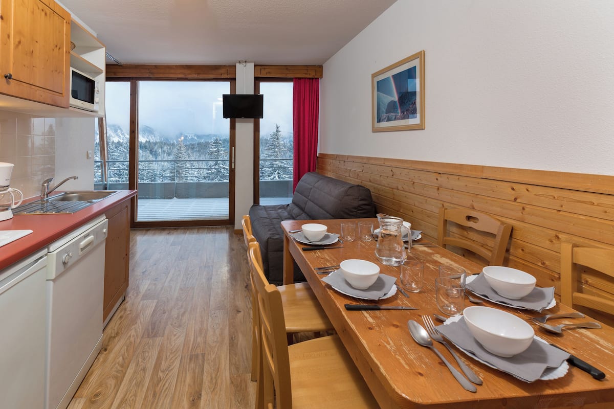 Un coin repas avec une table en bois dressée pour quatre personnes, un canapé et une cuisine équipée d’un four à micro-ondes.