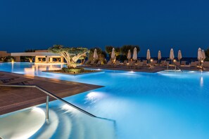 A nighttime swimming pool area with lounge chairs and umbrellas, surrounded by trees and a building.