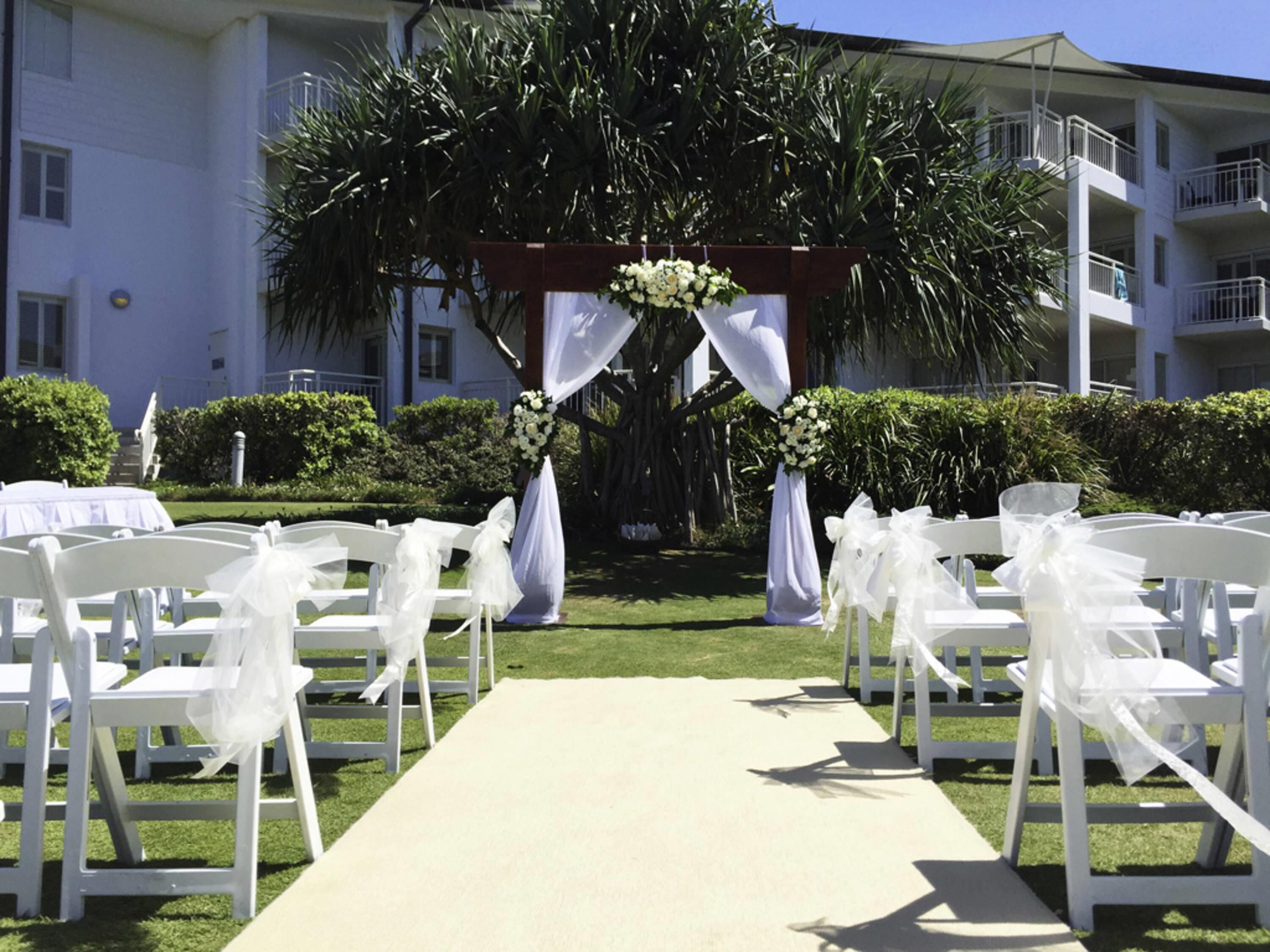 A wedding aisle with white chairs and a white carpet, leading to a decorated archway with white flowers and drapes.