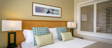 A hotel room with a bed, two bedside tables, a wall-mounted air conditioner, a window with blinds, and a framed picture above the bed.