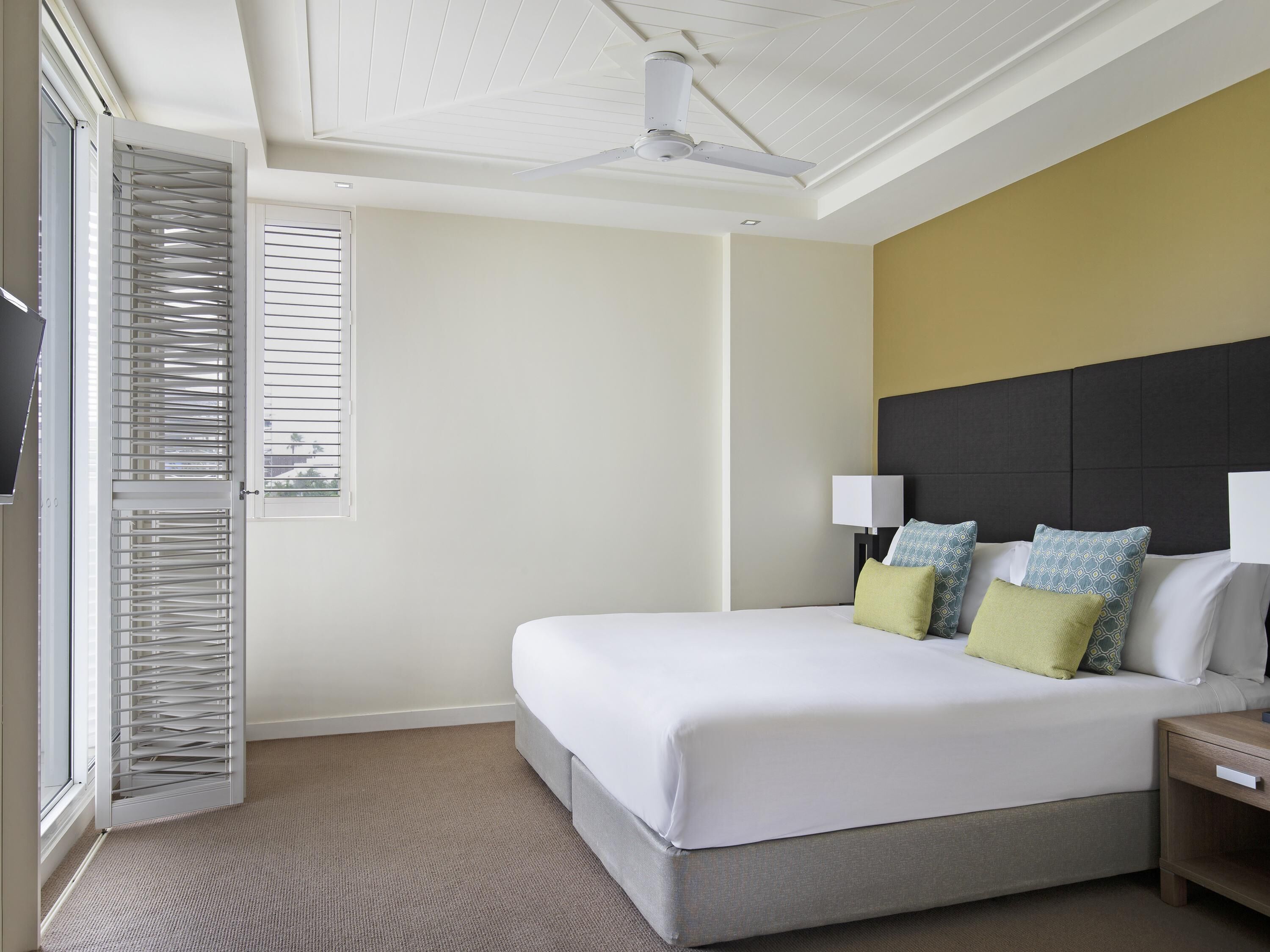 A hotel room with a bed, a nightstand, a ceiling fan, and a window with blinds.