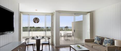 A spacious living area with a balcony, a flat-screen TV, a dining table, and a sofa with decorative pillows.