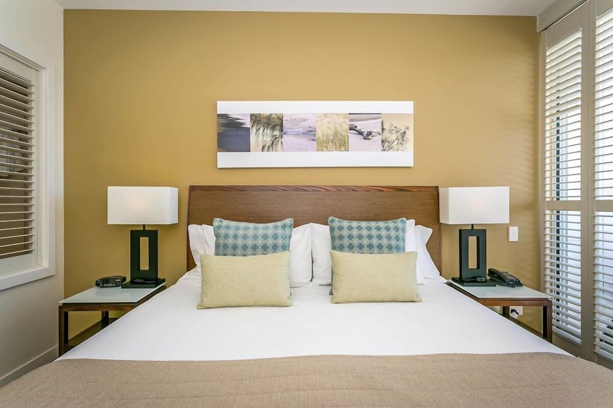 Quarto de hotel com cama grande, duas mesas de cabeceira com luminárias, cabeceira e obra de arte pendurada na parede.