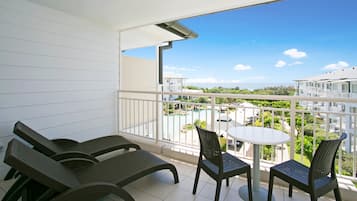 Een balkon met een witte reling, twee lounge stoelen, een kleine tafel en uitzicht op een zwembad en gebouwen.