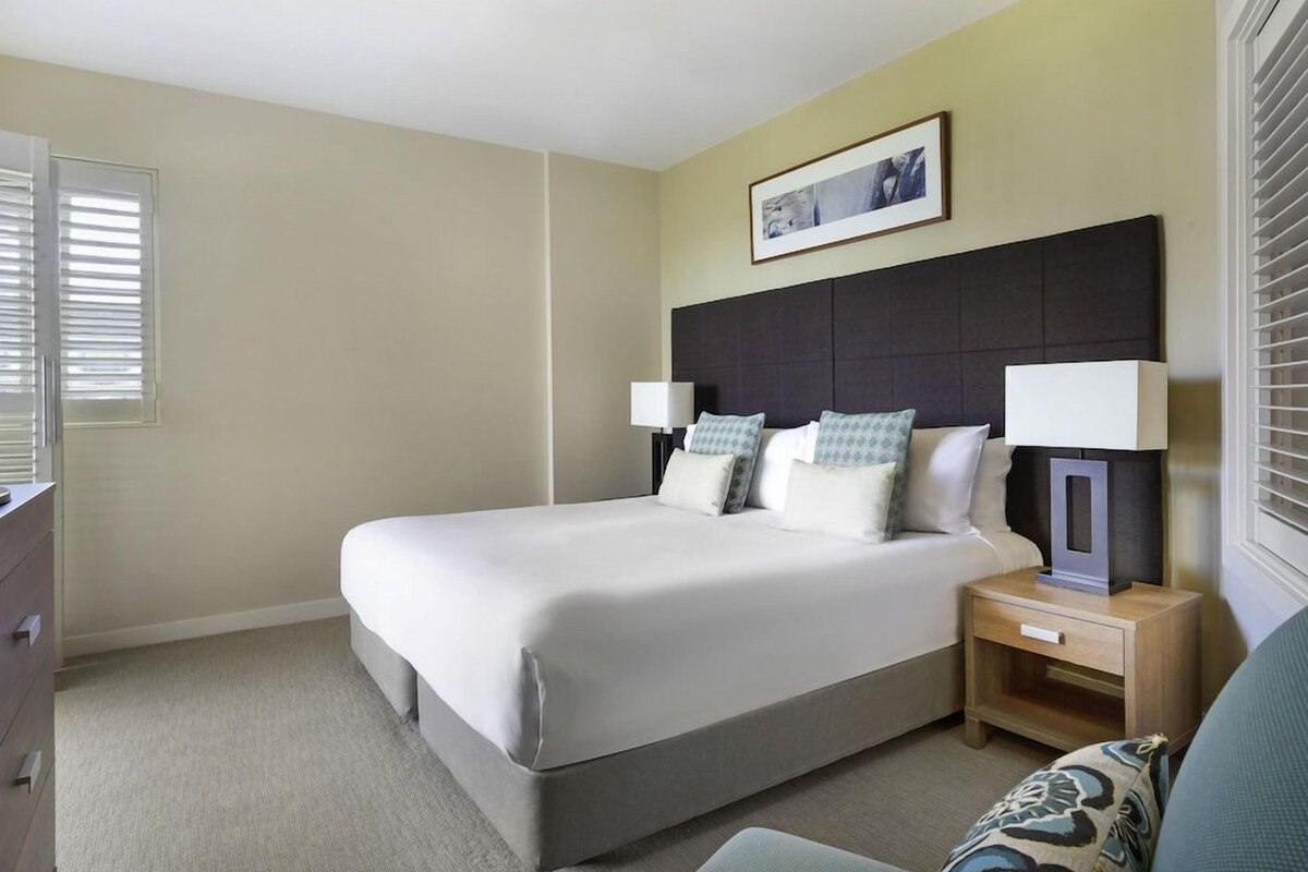 Quarto de hotel com cama, mesas de cabeceira, cômoda e janela com persianas.