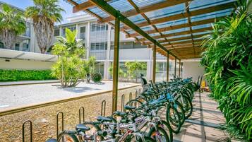 A covered bike parking area with rows of bicycles, palm trees, and a building in the background.
