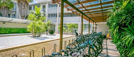 A covered bike parking area with rows of bicycles, palm trees, and a building in the background.