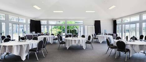 A conference room with multiple tables and chairs, large windows, and a high ceiling.
