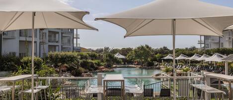A poolside area with lounge chairs and umbrellas, a pool, and a view of a building with balconies.