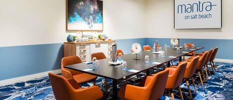 A conference room with a long table, orange chairs, a projector screen, and a large abstract painting.