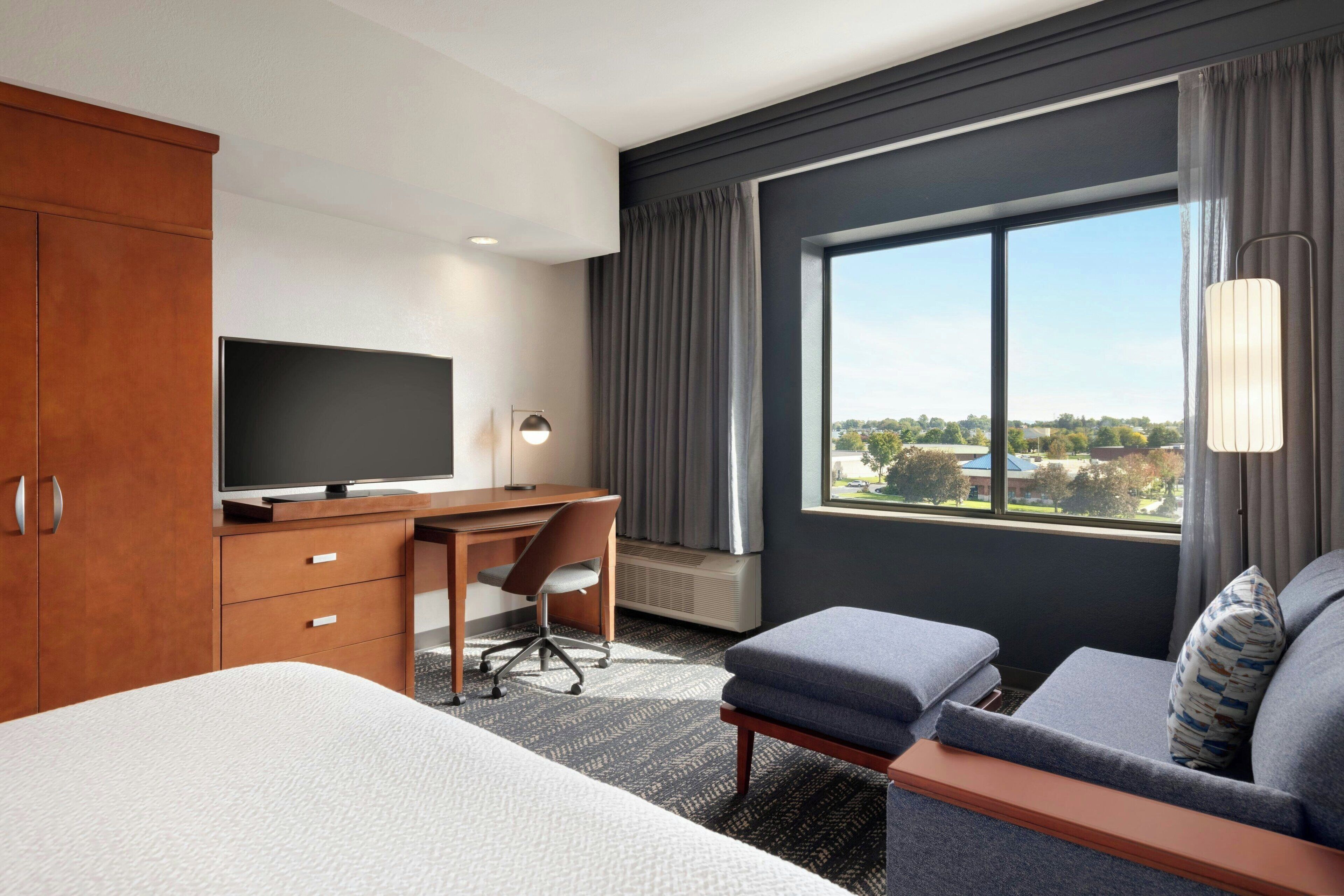 A hotel room with a bed, a desk with a television, a chair, a sofa, and a large window offering a view of the city.
