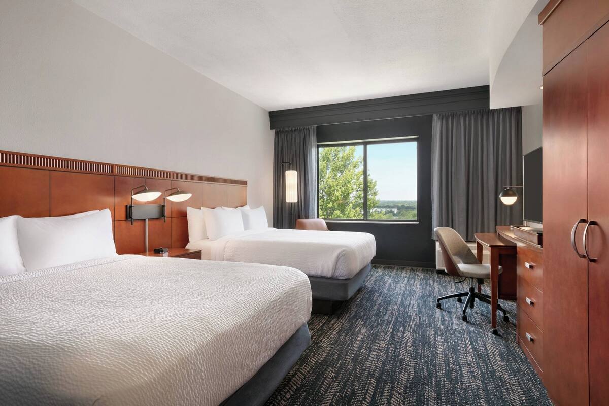 A hotel room with two beds, a desk, a chair, and a view of the outdoors.