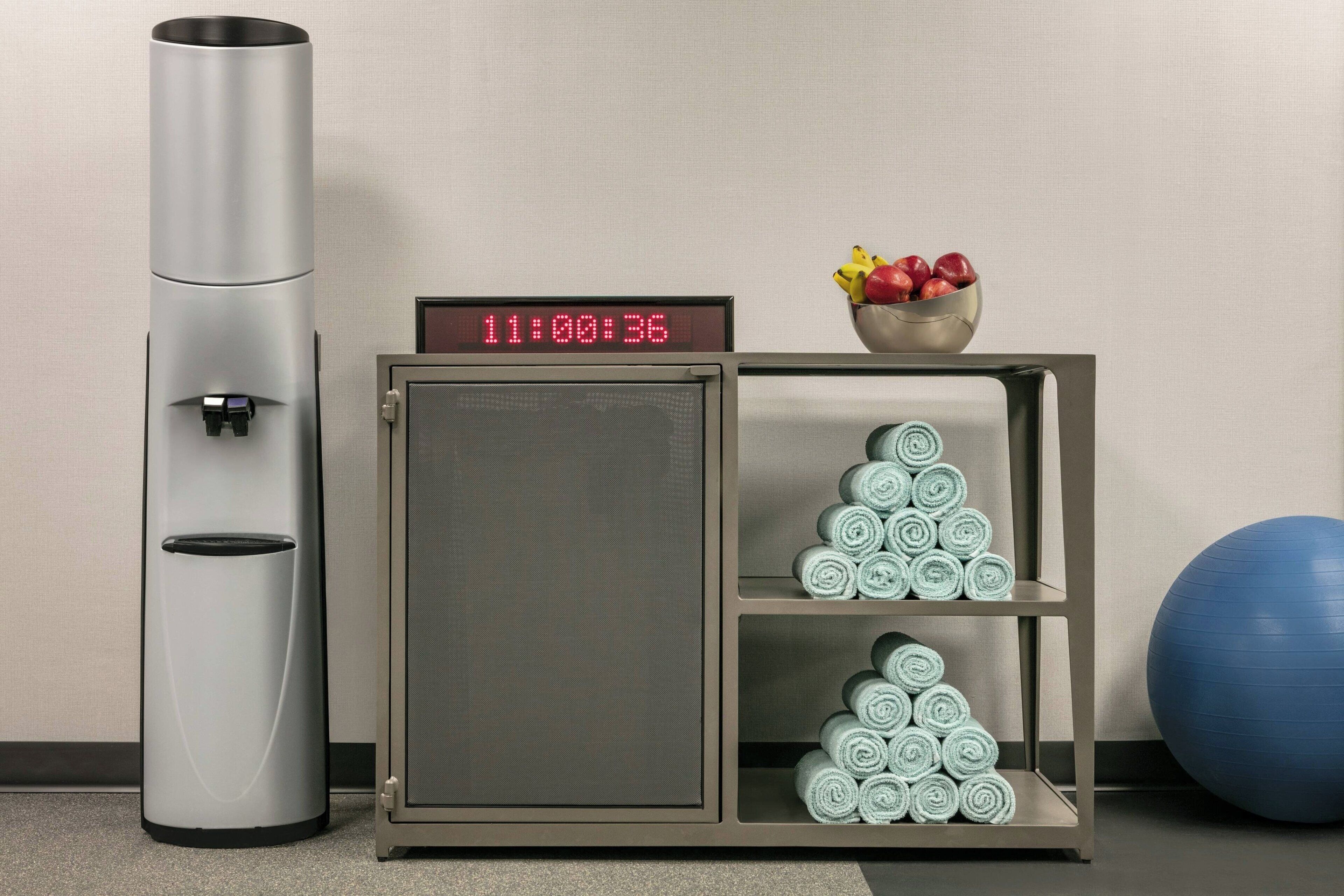 A compact room with a digital clock, a waste bin, a small cabinet with towels, and a blue exercise ball.