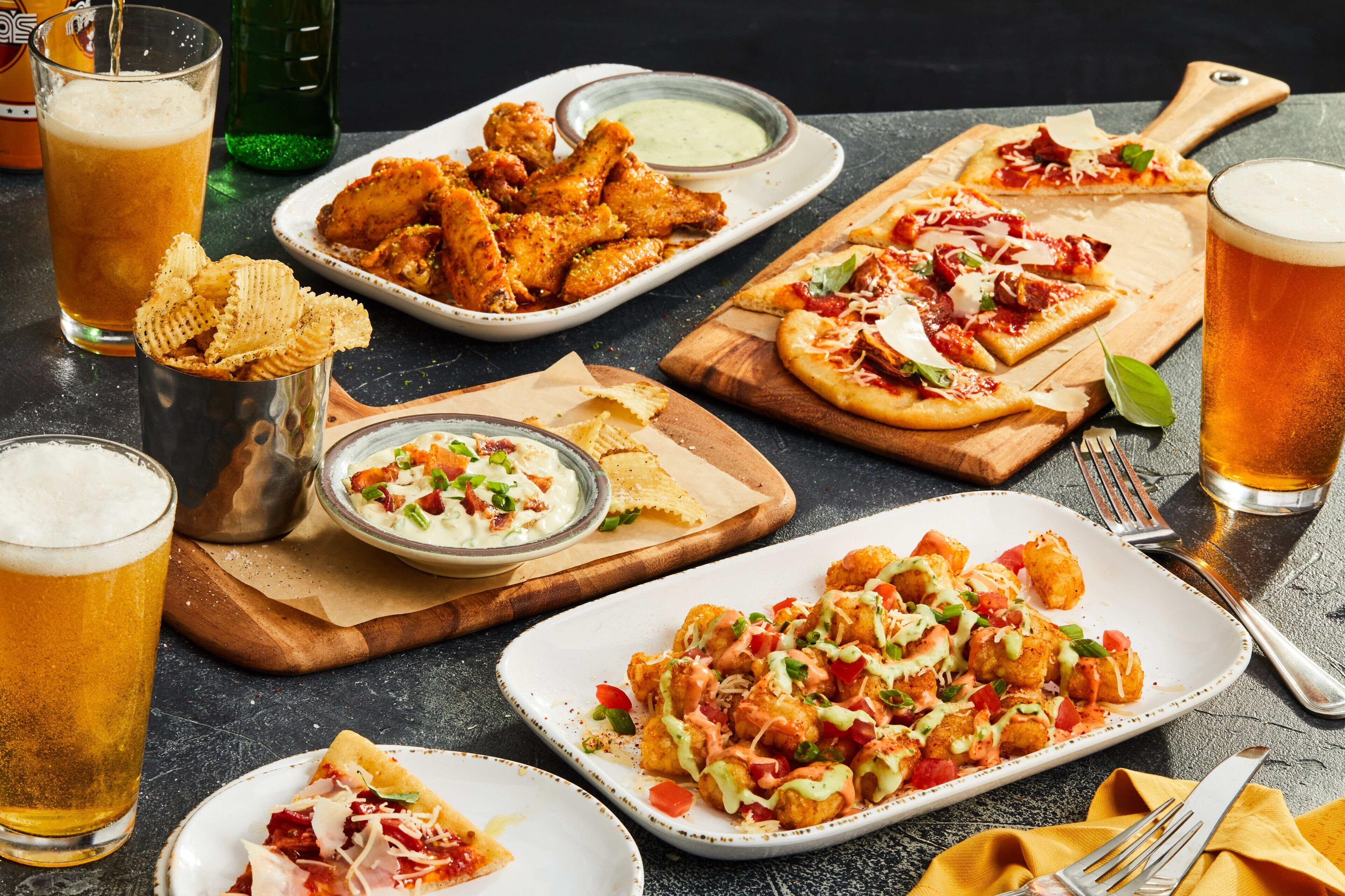 A variety of dishes including wings, pizza, and tater tots, along with glasses of beer.