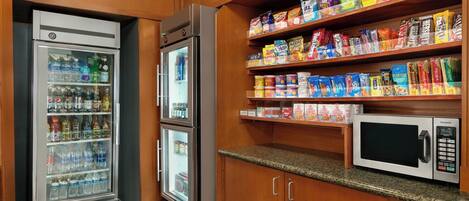 A kitchenette with a refrigerator, a microwave, and shelves stocked with snacks and beverages.