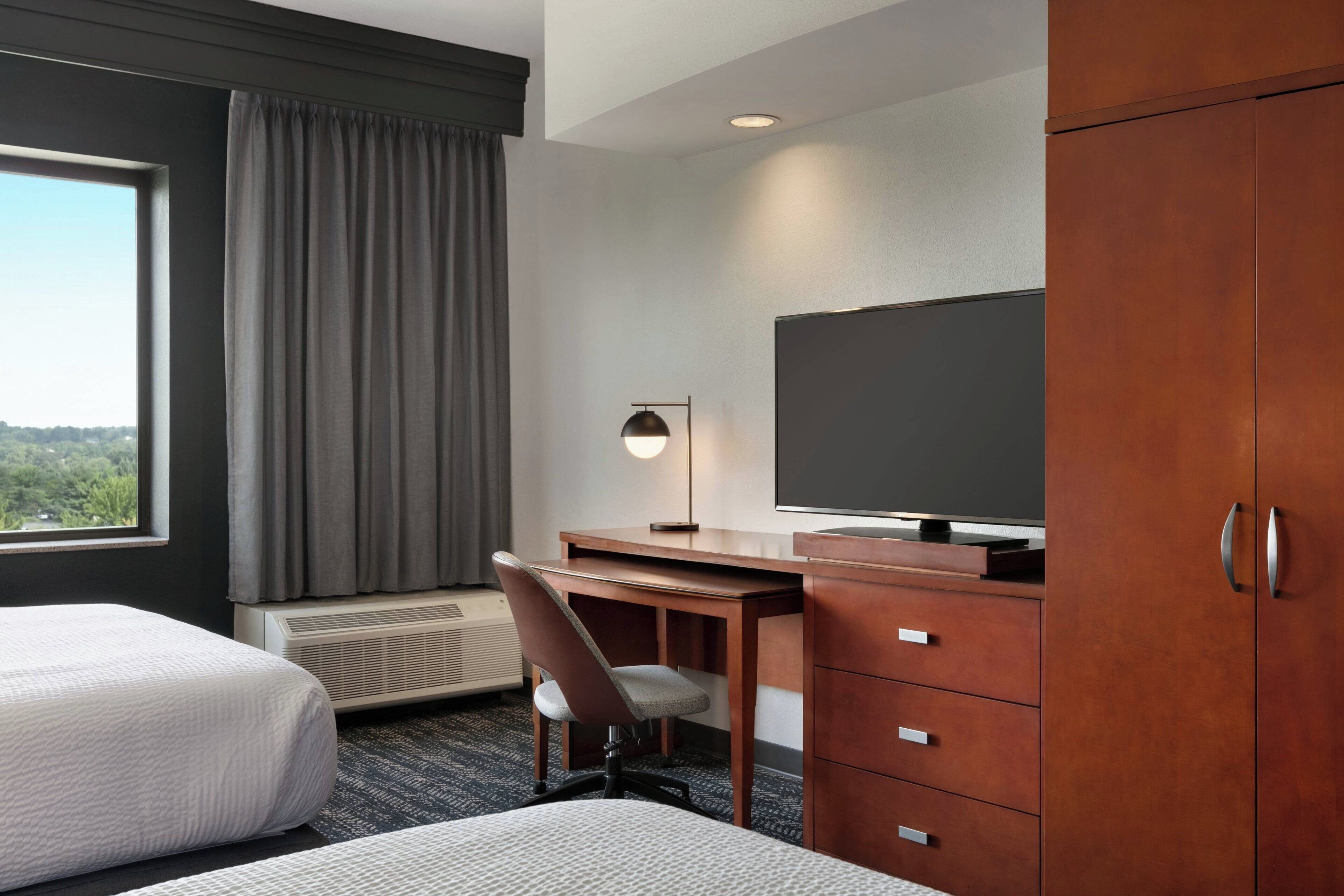 A hotel room with two beds, a desk, a chair, a television, a wardrobe, and a view of the outdoors.