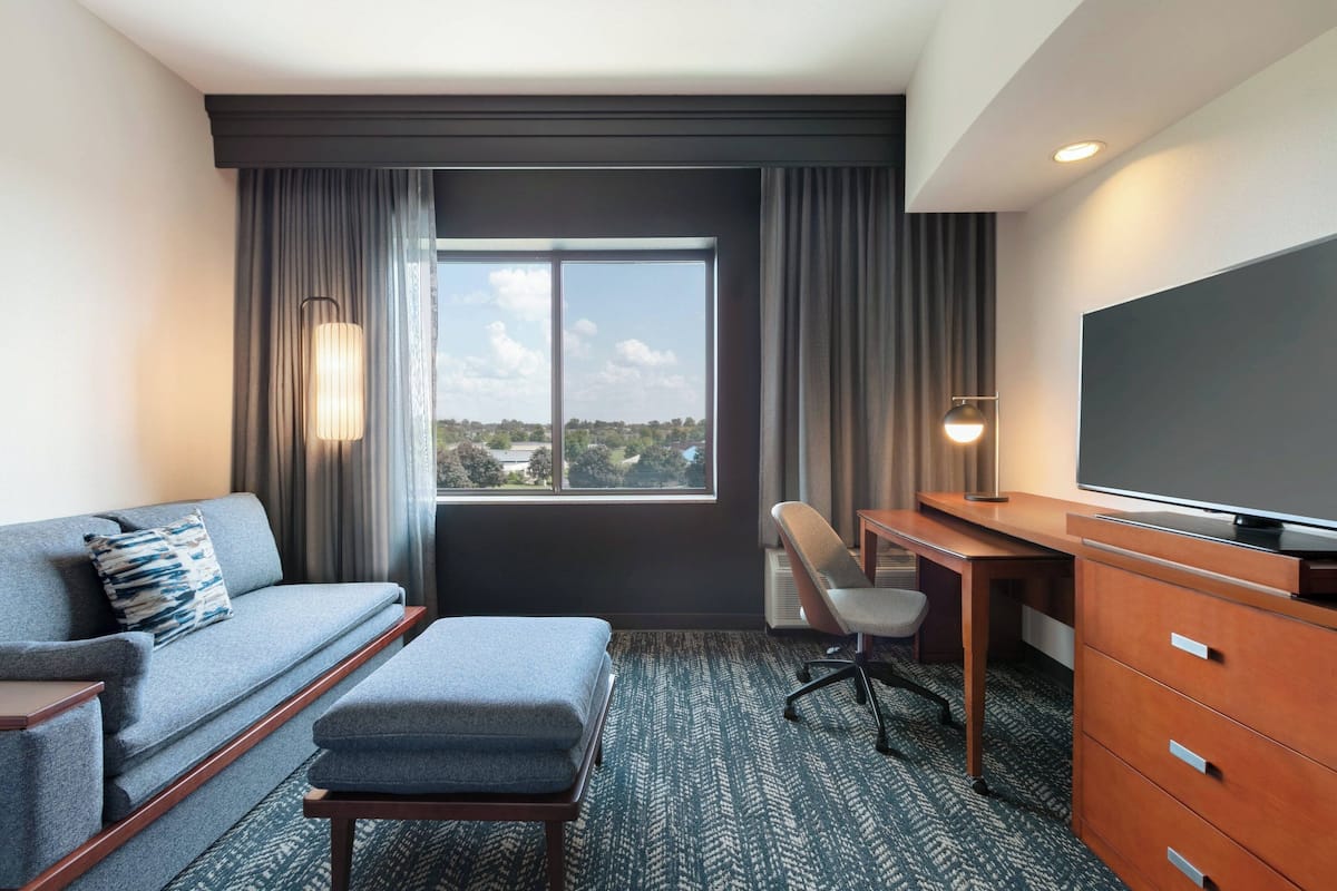 A hotel room with a sofa, a desk, a chair, a television, and a large window with curtains.