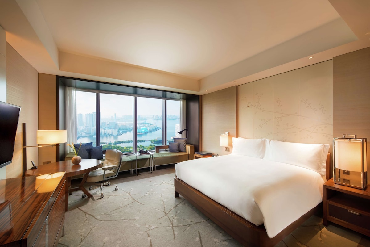 A hotel room with a large bed, a desk, a chair, a TV, and a view of a cityscape through large windows.
