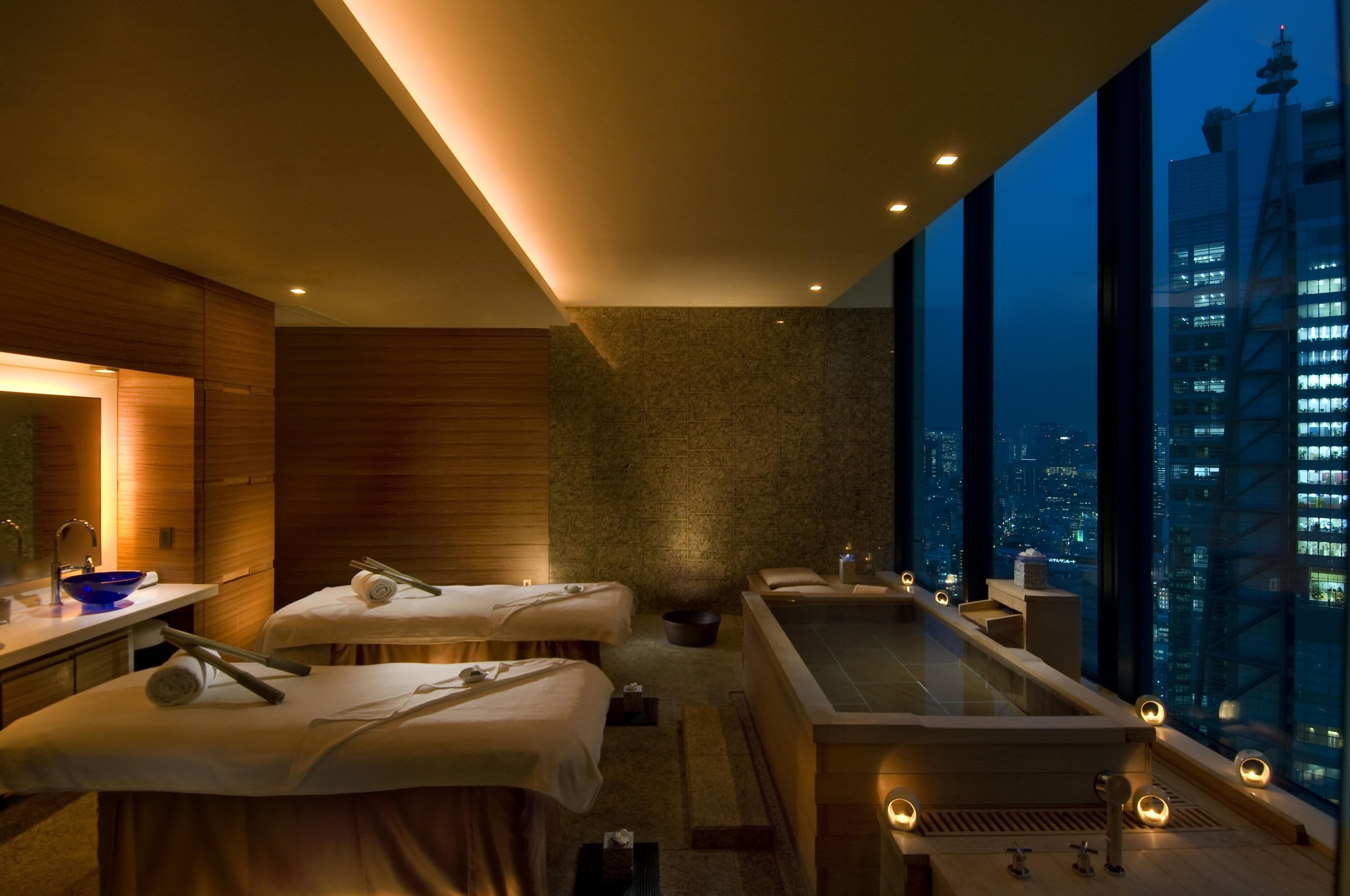Une salle de spa moderne dotée d’une grande baignoire, de deux tables de massage et offrant une vue sur la ville.