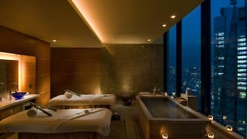 Une salle de spa moderne dotée d’une grande baignoire, de deux tables de massage et offrant une vue sur la ville.