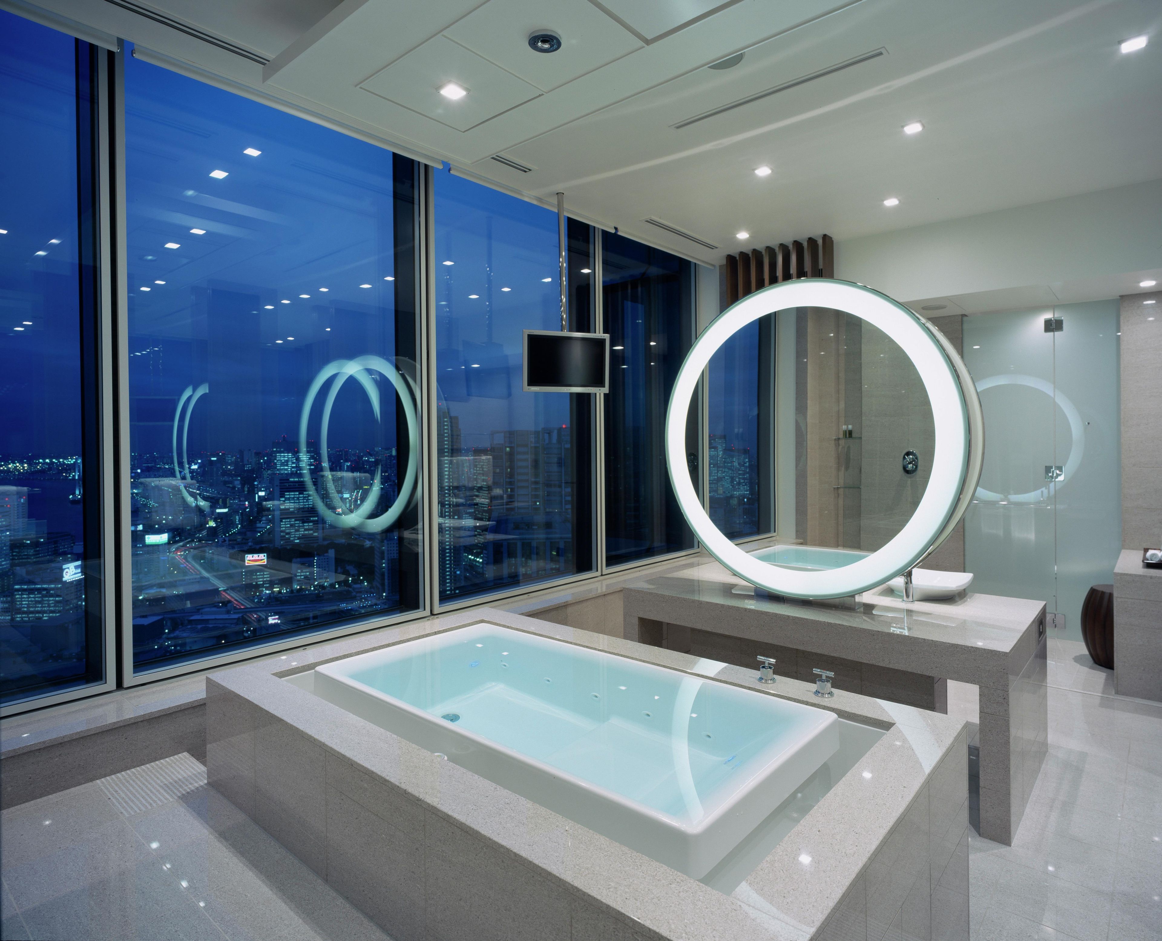 A modern bathroom with a large bathtub, a round mirror, and a view of a cityscape through large windows.