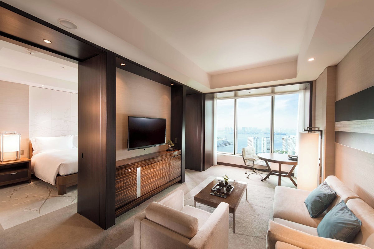 A modern hotel room with a large flat-screen TV, a sofa, a coffee table, and a view of the city through large windows.