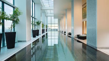 A long indoor swimming pool with clear glass walls, potted plants, and seating areas along the sides.