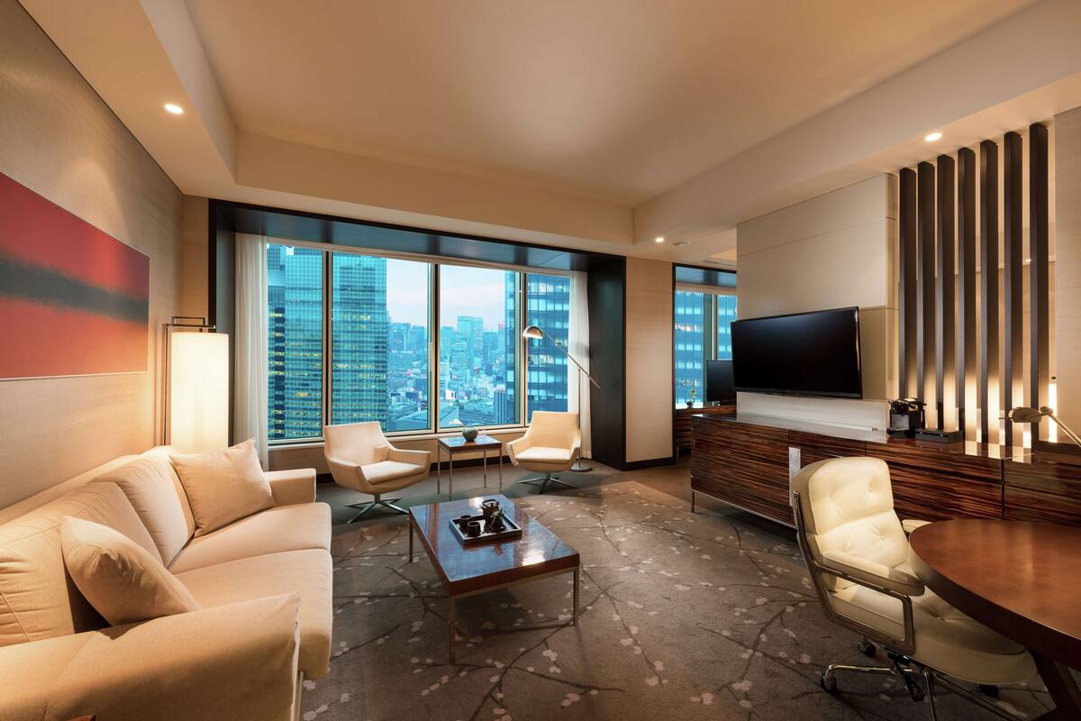 Una habitación de hotel moderna con un ventanal grande, un televisor de pantalla plana, un sofá, una mesa de centro y una mesa de comedor.