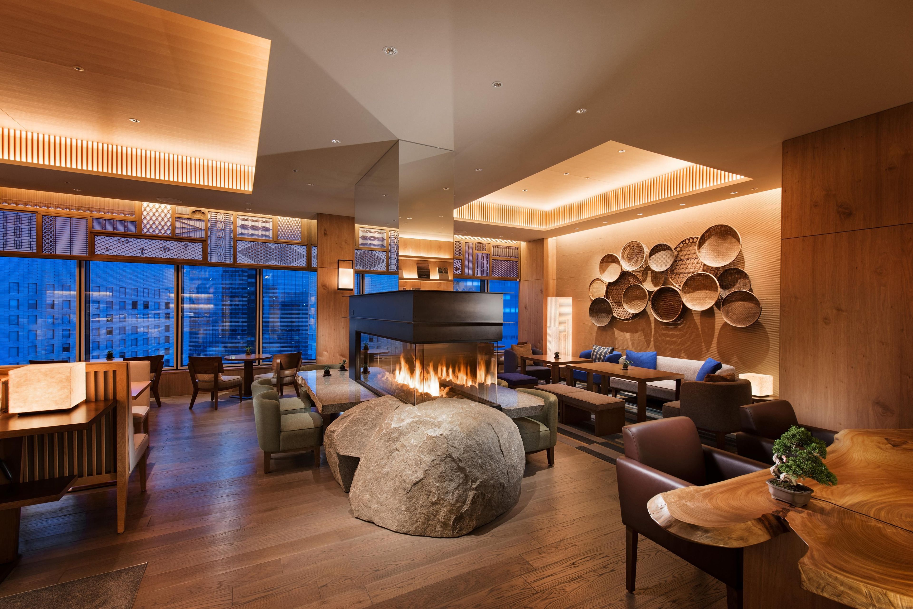 A modern interior with a central fire feature, wooden furniture, and a view of the cityscape through large windows.