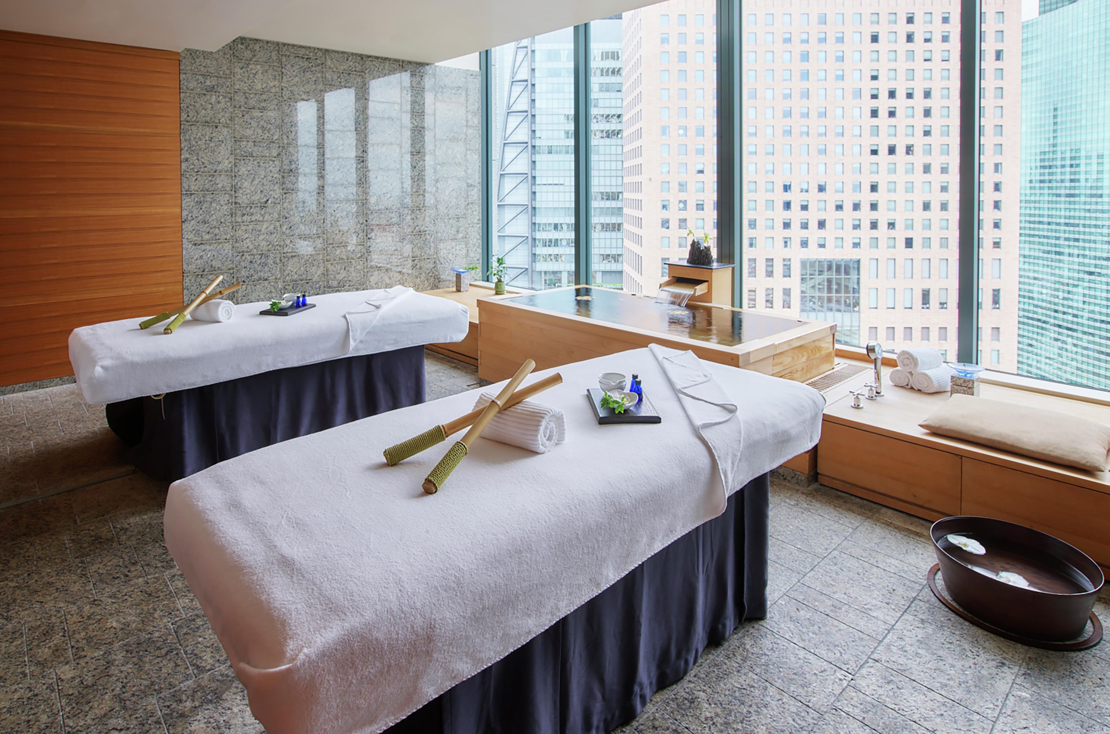 Un cuarto de spa con dos camillas de masaje, un lavamanos pequeño y vistas a la ciudad a través de amplias ventanas.