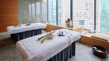 Un cuarto de spa con dos camillas de masaje, un lavamanos pequeño y vistas a la ciudad a través de amplias ventanas.