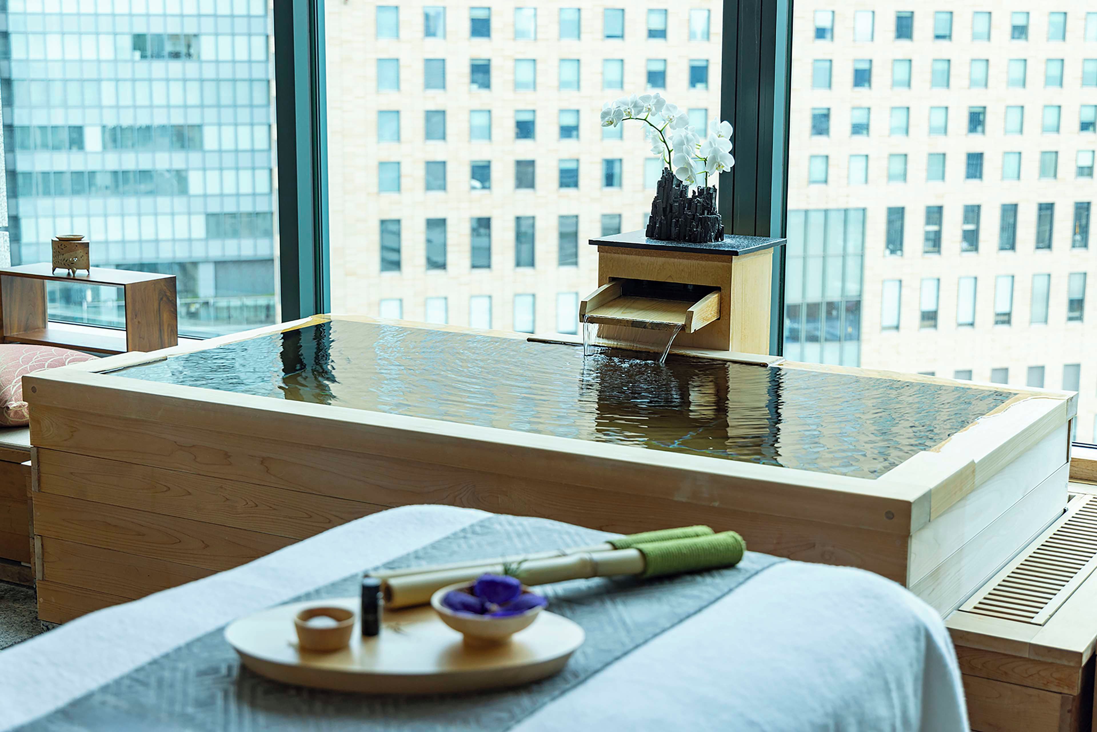 A modern spa room with a wooden bathtub, a small wooden table with a flower, and a window with a city view.