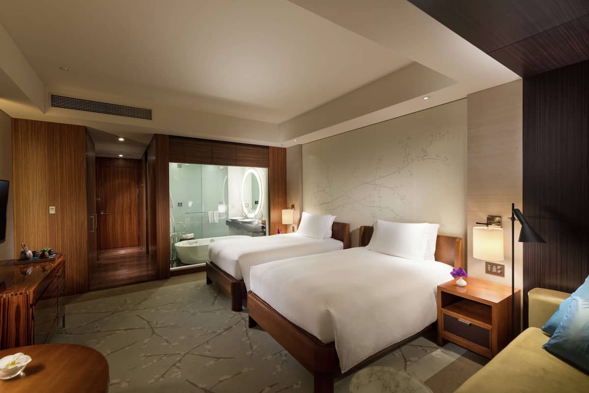 Habitación de hotel con dos camas, un escritorio de madera, un baño con mampara de vidrio y un espejo grande.