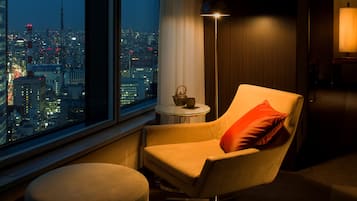 A modern hotel room with a large window offering a city view, a comfortable armchair with a red pillow, a small round table with a lamp, and a decorative item on the table.