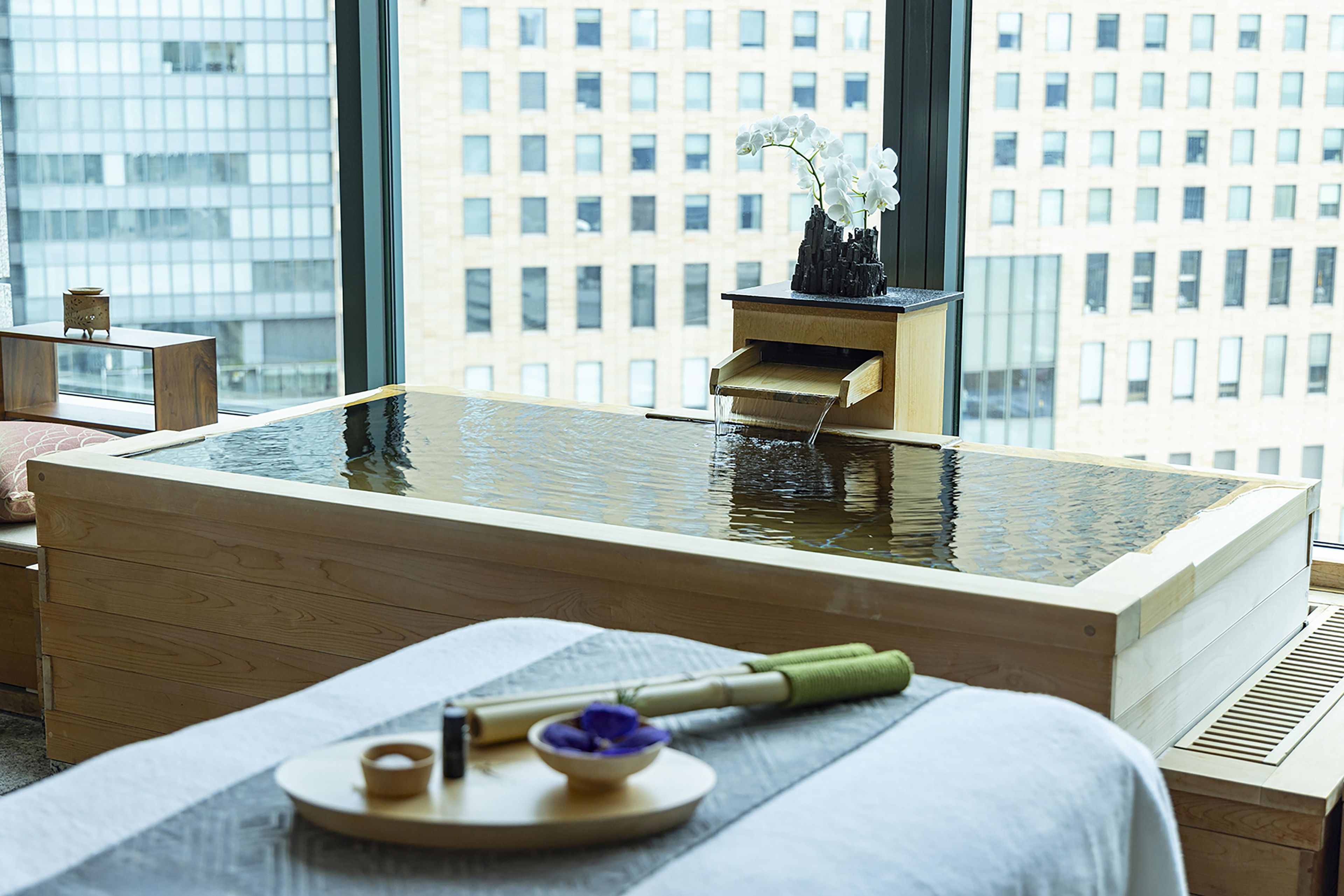 Una moderna stanza spa con una vasca da bagno in legno, un piccolo tavolino con una ciotola e una spazzola, e una finestra che si affaccia su un panorama urbano.