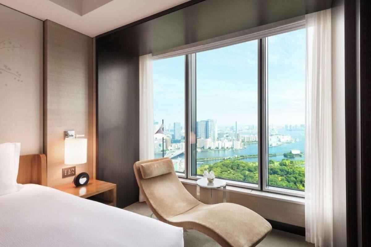 Habitación de hotel con cama, sillón, mesita de noche con lámpara y ventana grande con vistas a la ciudad.